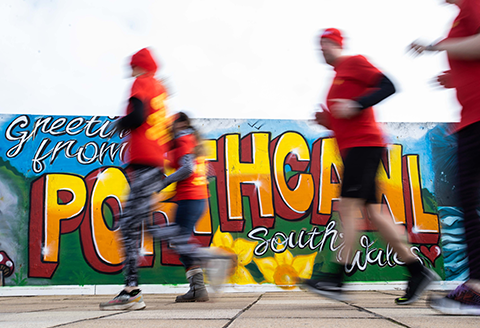 People running in red Ogi hats and t shirts in front of a wall with graffiti art that reads 