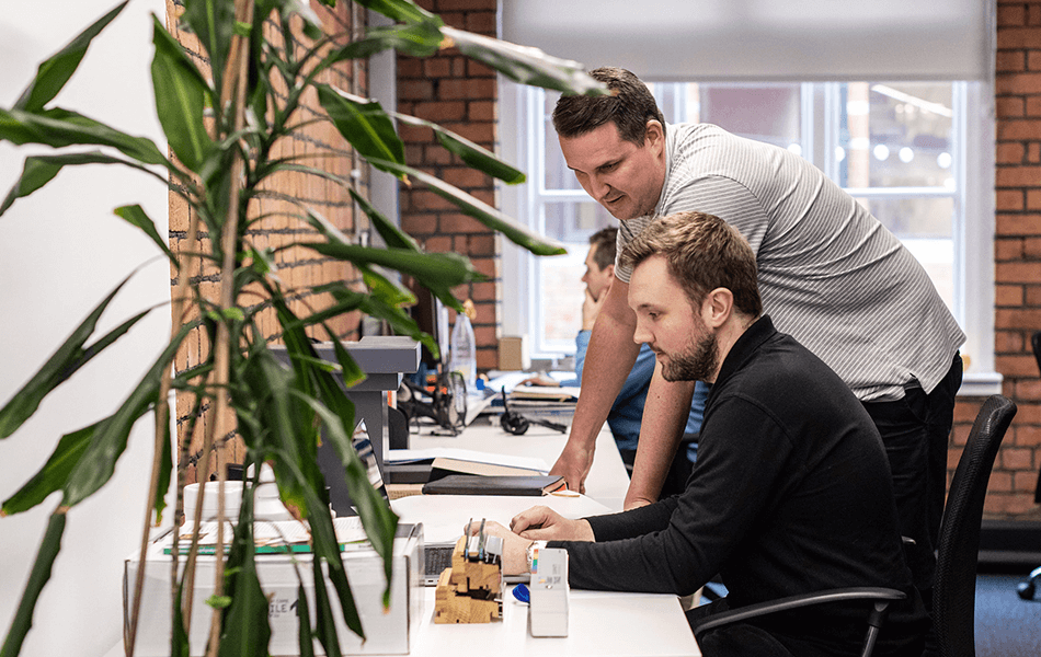 Two office workers looking at a computer. One is standing while the other is sitting.